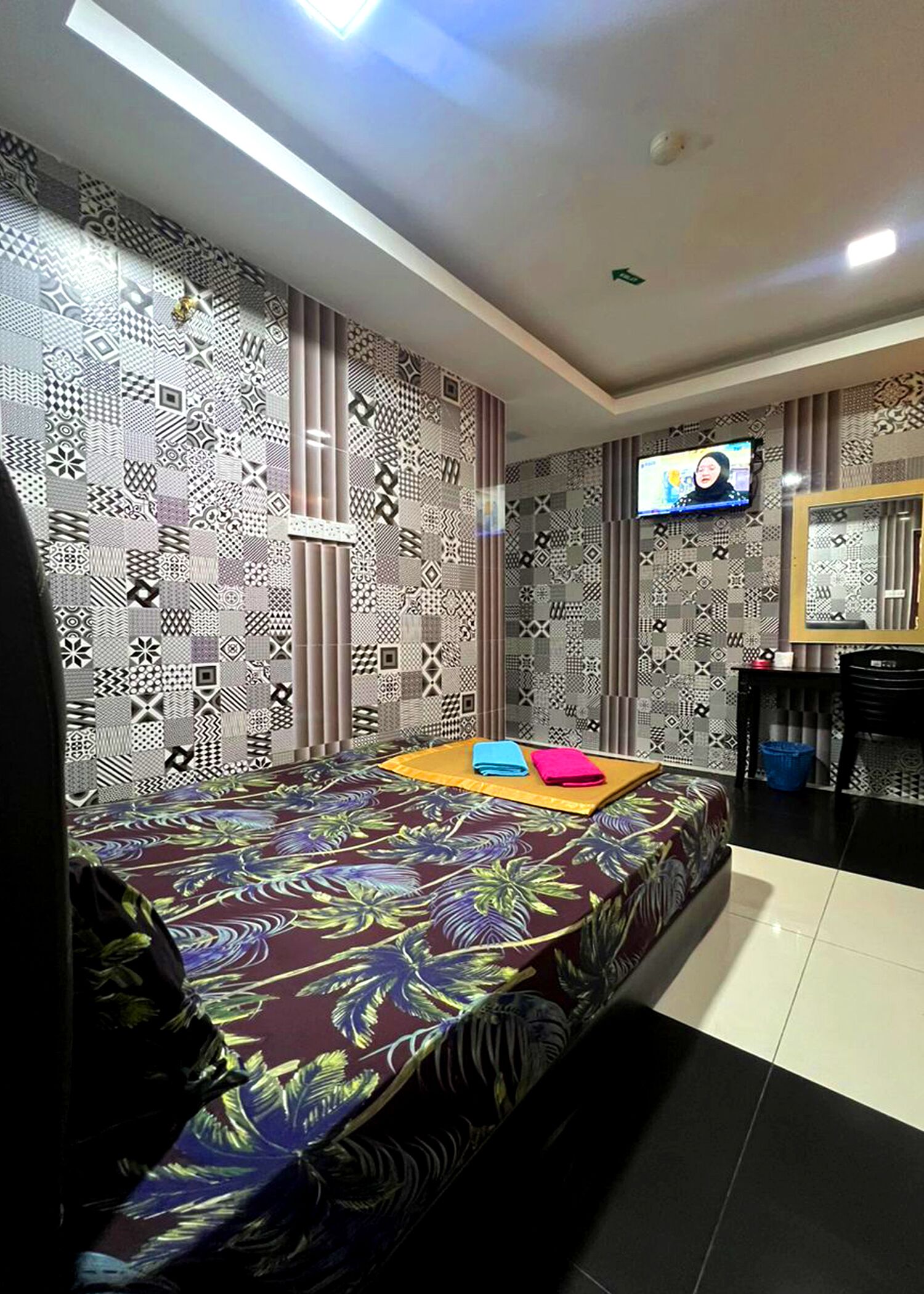 Habitación de hotel con pared estampada, cama con colcha de motivos tropicales, televisor de pantalla plana y una pequeña mesa con una silla.