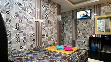 Habitación de hotel con pared estampada, cama con colcha de motivos tropicales, televisor de pantalla plana y una pequeña mesa con una silla.