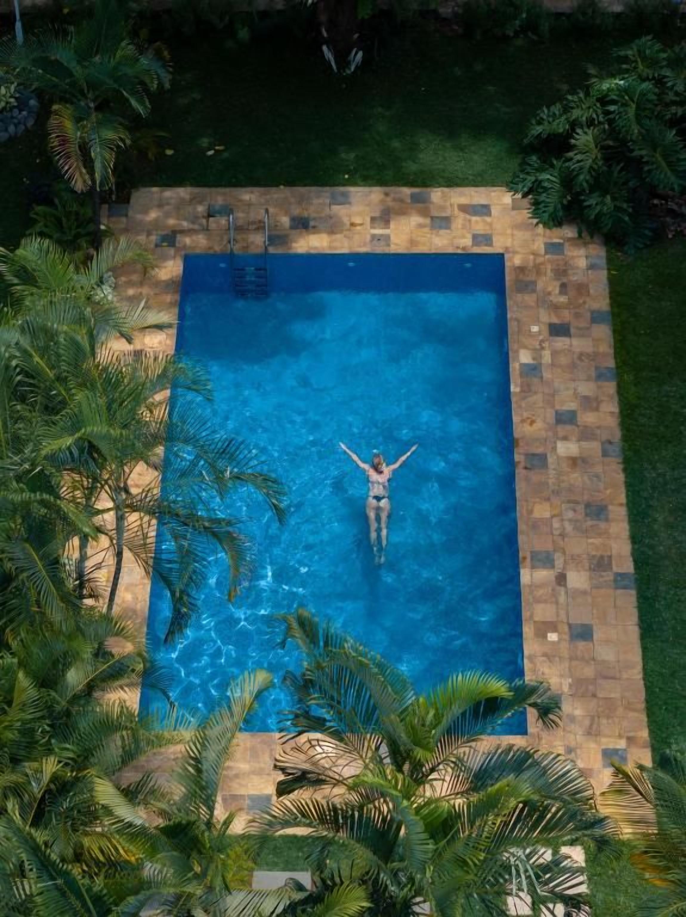 A rectangular swimming pool with a tiled border, surrounded by palm trees and greenery.