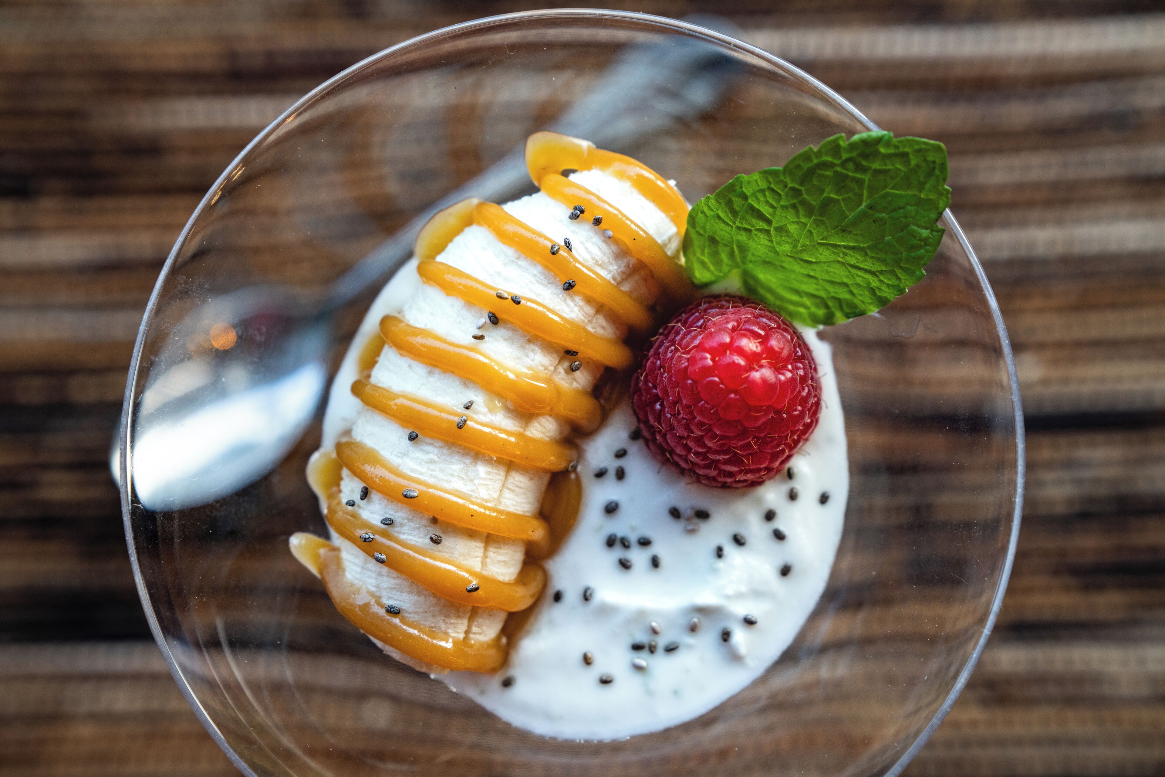 A dessert in a clear glass bowl with sliced bananas, a strawberry, and a mint leaf on a wooden surface.