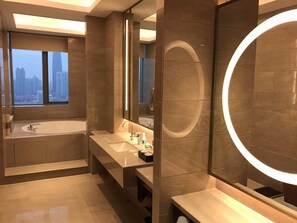 Une salle de bain moderne dotée d’une grande baignoire, d’un meuble-lavabo avec un miroir et offrant une vue sur le paysage urbain grâce à de grandes fenêtres.
