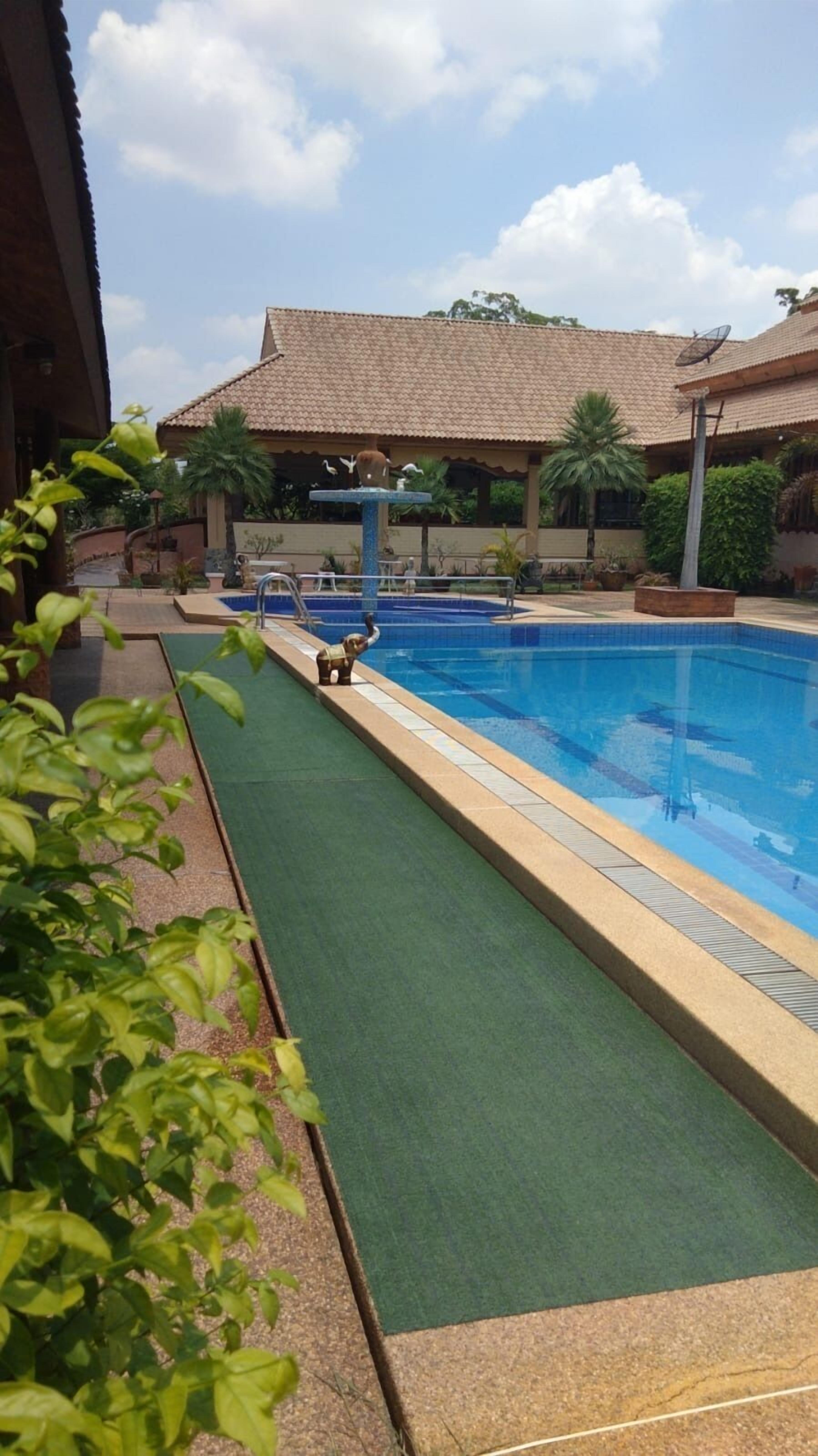 A swimming pool with a tiled deck, surrounded by greenery and a building with a tiled roof.