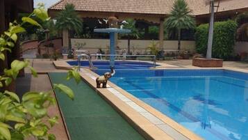 A swimming pool with a tiled deck, surrounded by greenery and a building with a tiled roof.