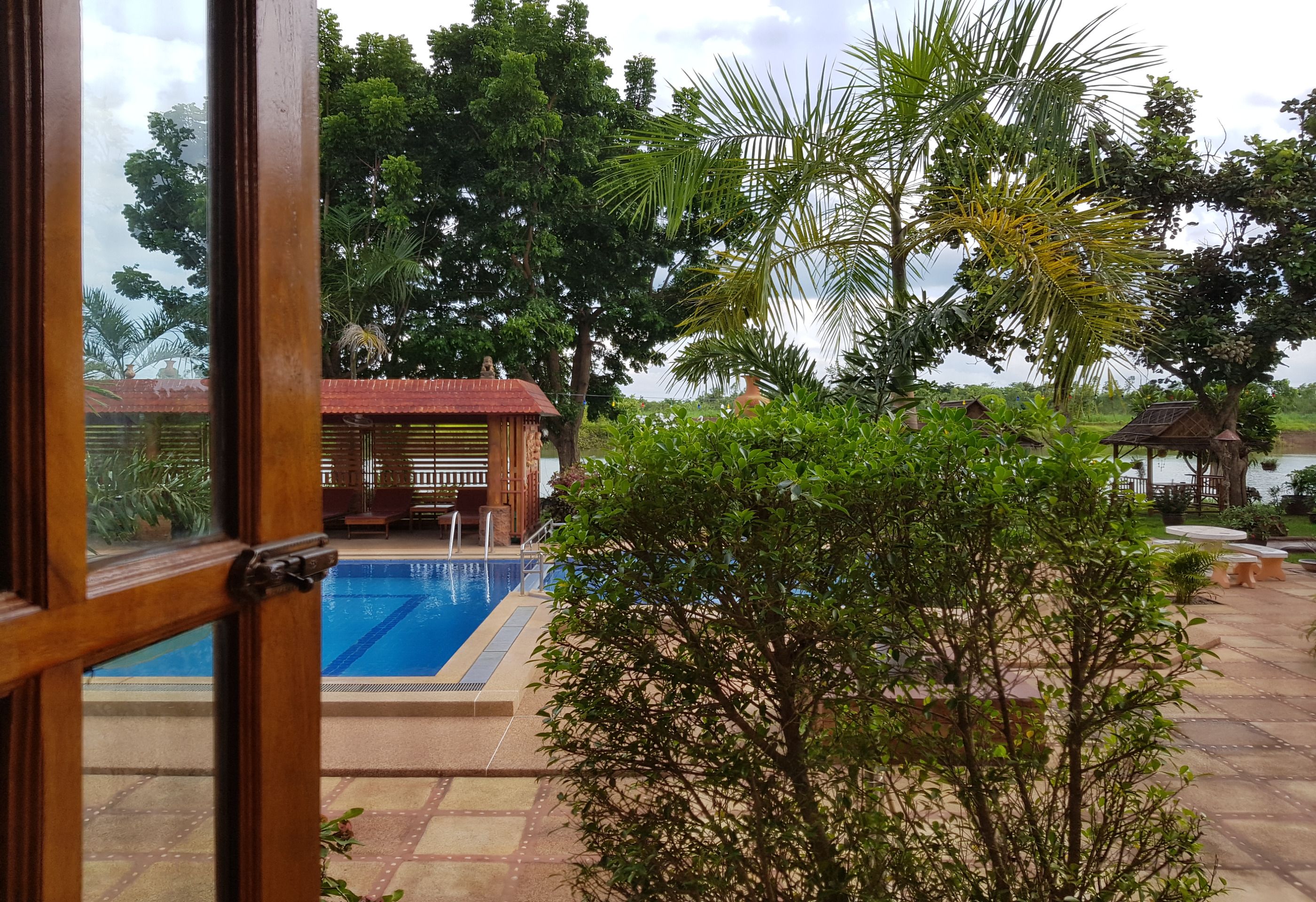 Uma área de piscina com deck de madeira, um pequeno edifício com telhado vermelho e vegetação exuberante.