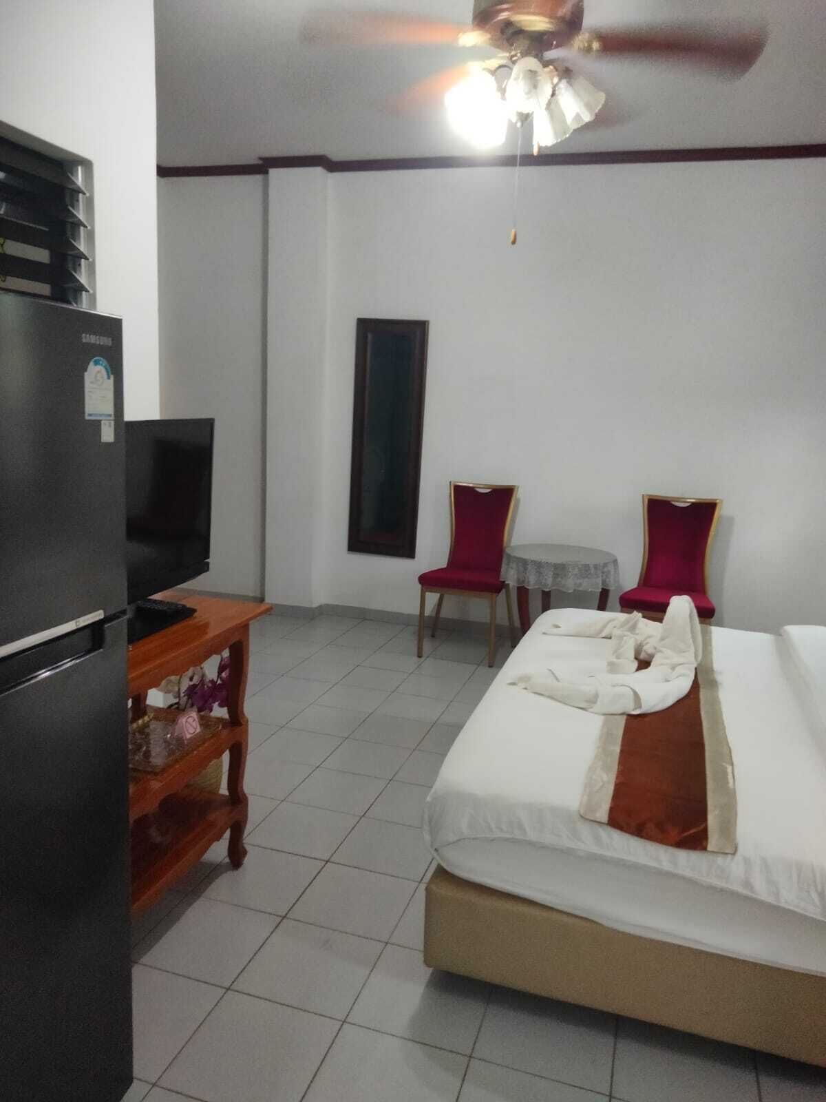 A hotel room with a bed, a desk, two chairs, a refrigerator, and a ceiling fan.