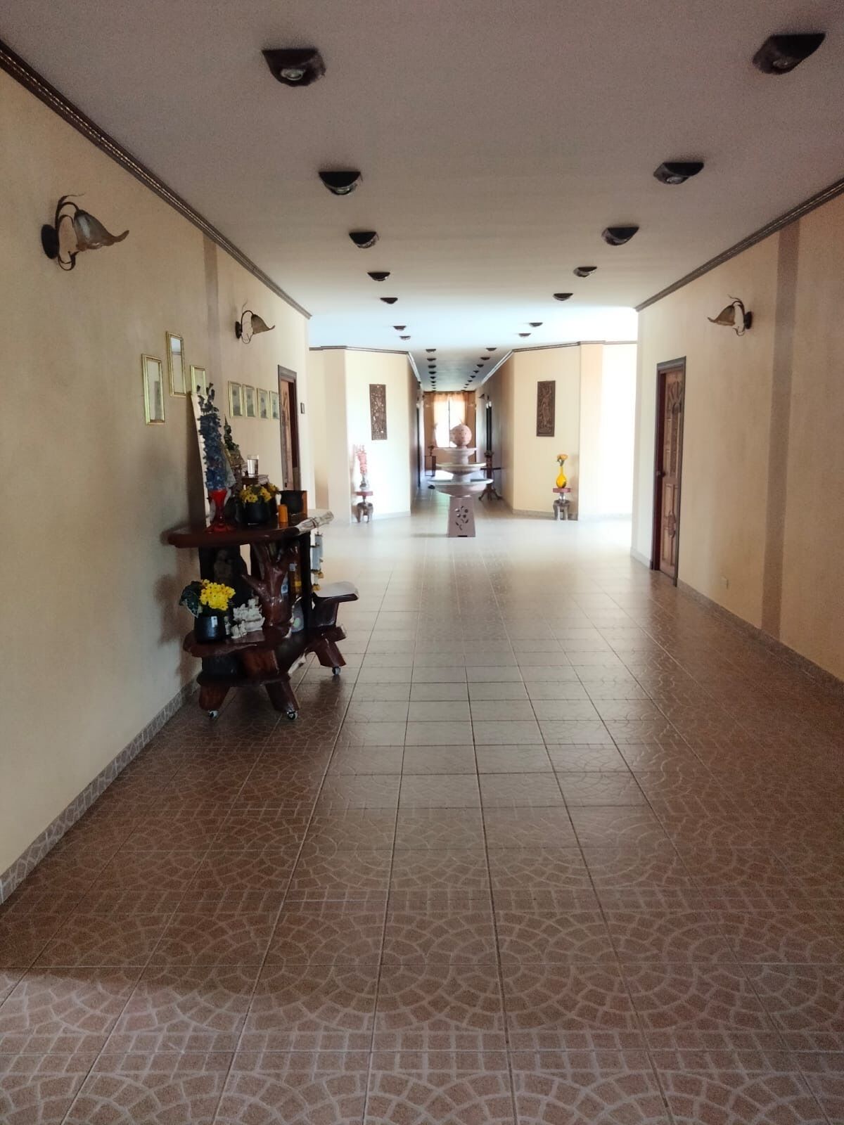 A long, tiled hallway with a central fountain, flanked by walls with framed pictures and wall-mounted lights.
