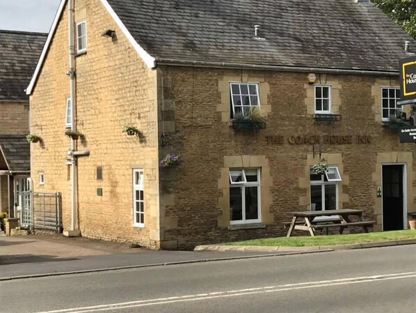Ein Steinhaus mit einem Schild, auf dem "The Coach House Inn" steht. Es gibt Fenster und eine Tür, und eine Bank vor dem Haus.