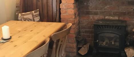 Una mesa de madera para comer con cuatro sillas, una chimenea de ladrillo con leña, y una pared de piedra.