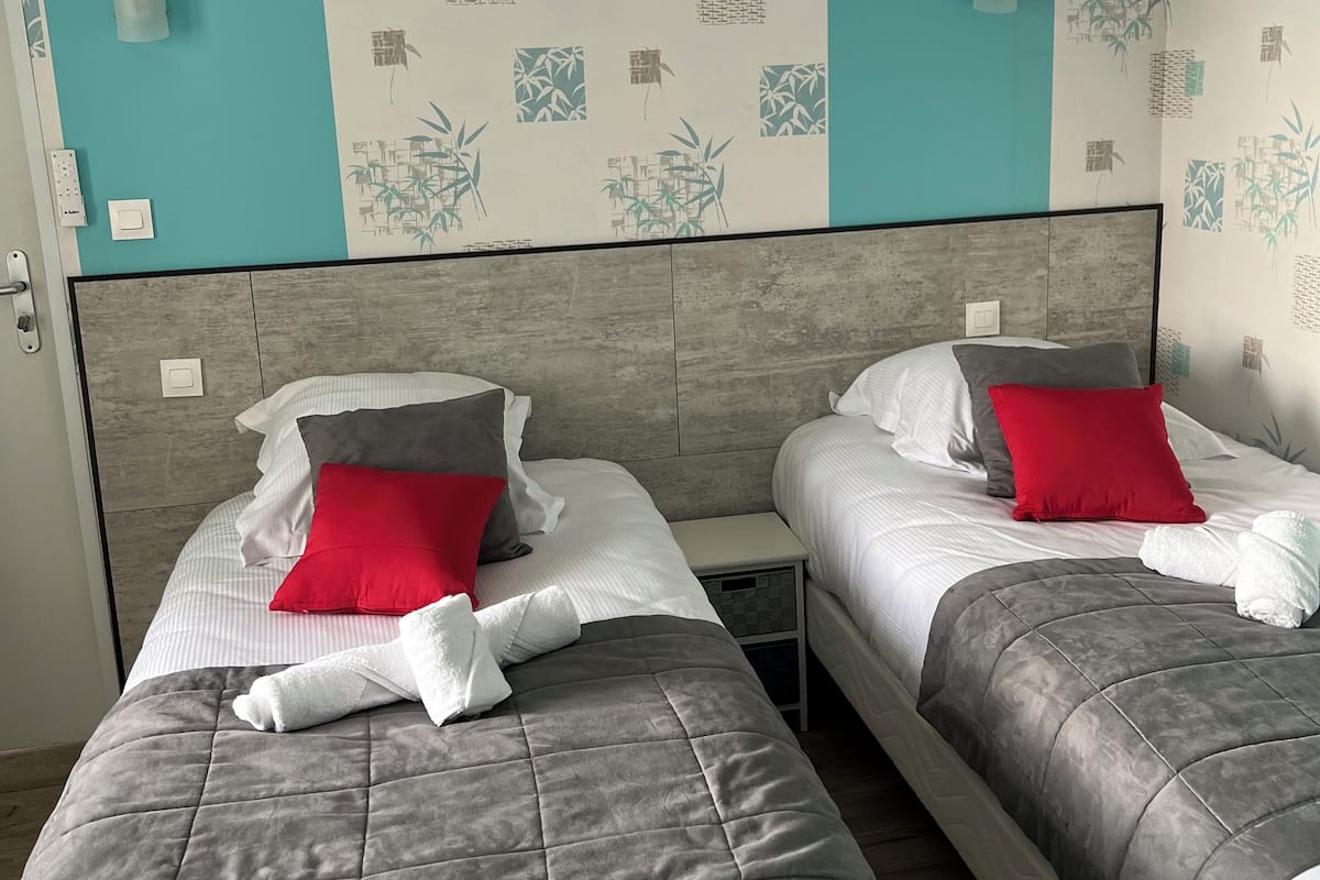 A hotel room with two beds, each with a grey quilted bedspread and pillows in red and white. The walls have a bamboo pattern wallpaper, and there are wall-mounted lights.