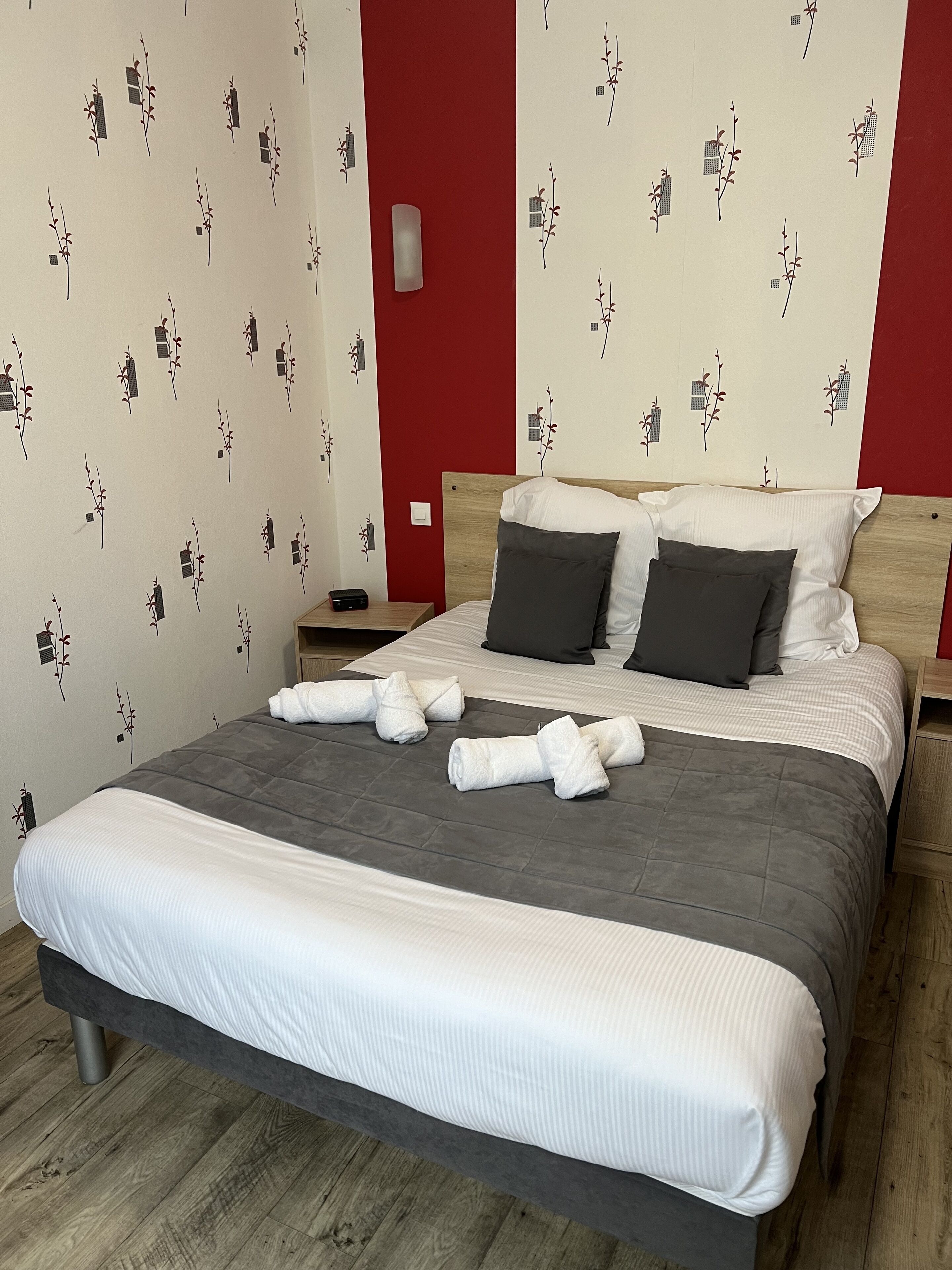 Un lit bien fait, avec une couette grise, deux oreillers foncés et une couverture blanche. Il y a deux tables de chevet, dont l’une est surmontée d’une lampe. Les murs sont recouverts d’un papier peint à motifs floraux.