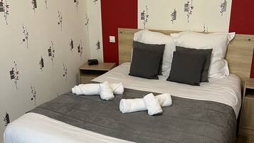 Un lit bien fait, avec une couette grise, deux oreillers foncés et une couverture blanche. Il y a deux tables de chevet, dont l’une est surmontée d’une lampe. Les murs sont recouverts d’un papier peint à motifs floraux.