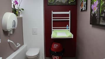 Un baño pequeño con inodoro, secamanos, lavabo, espejo y estante con una planta.