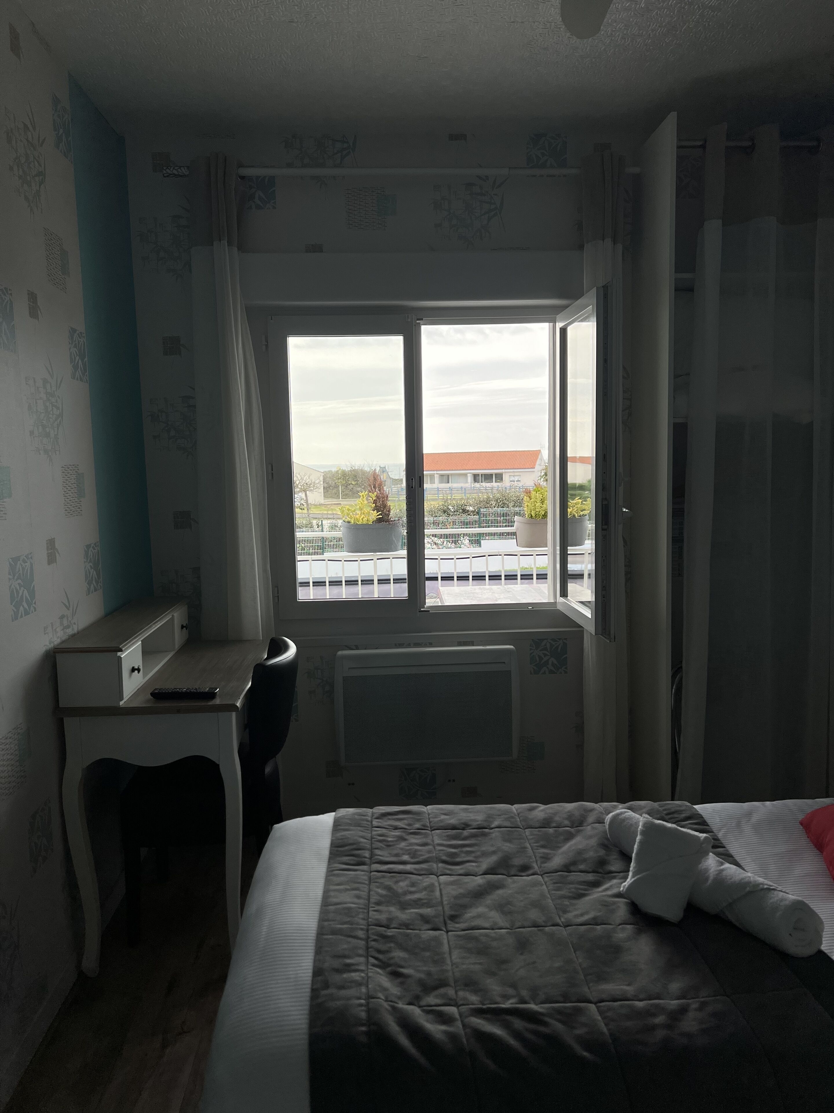 A hotel room with a bed, a desk, and a window with a view of buildings and a fence.