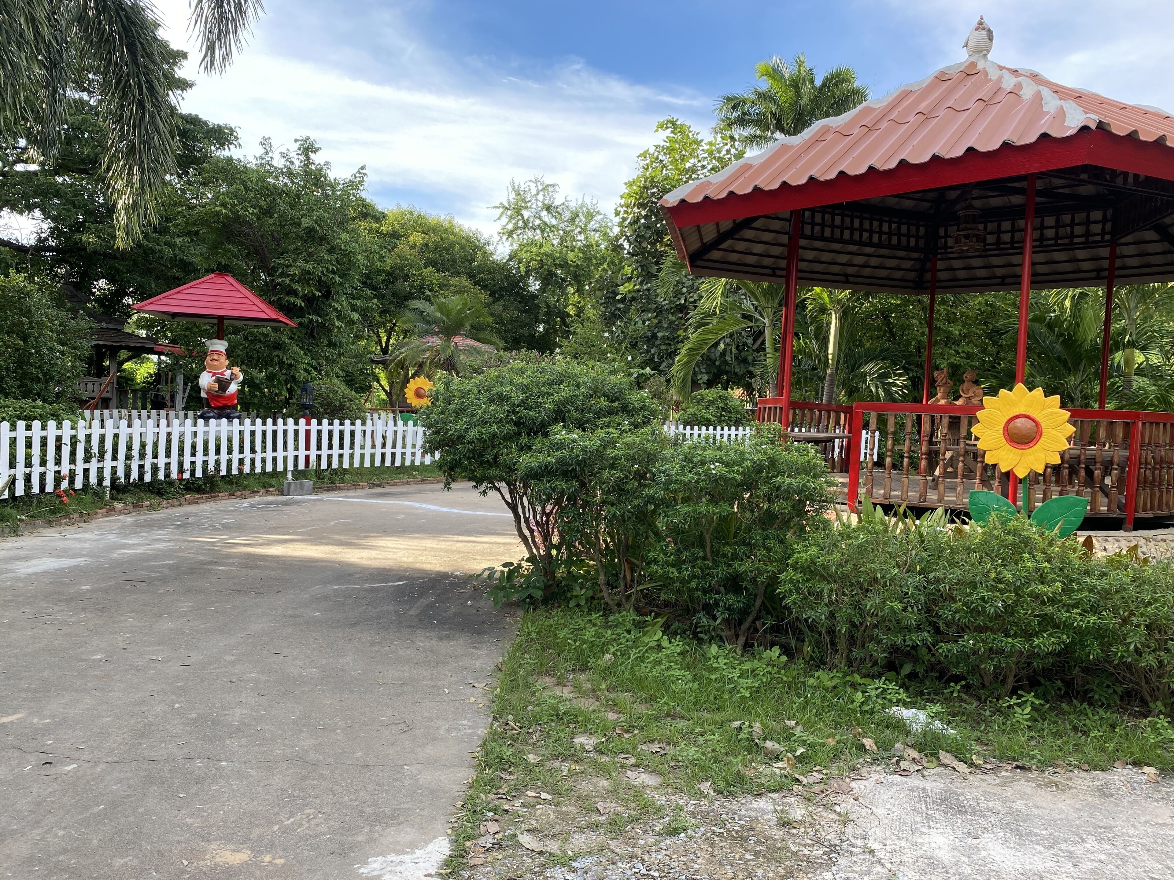 A gazebo with a red roof and a sunflower decoration, surrounded by greenery and a white picket fence.