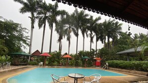 A swimming pool with two metal chairs and a table, surrounded by palm trees and covered structures. - Himawari Restaurant & Cottage (Khon Kaen)