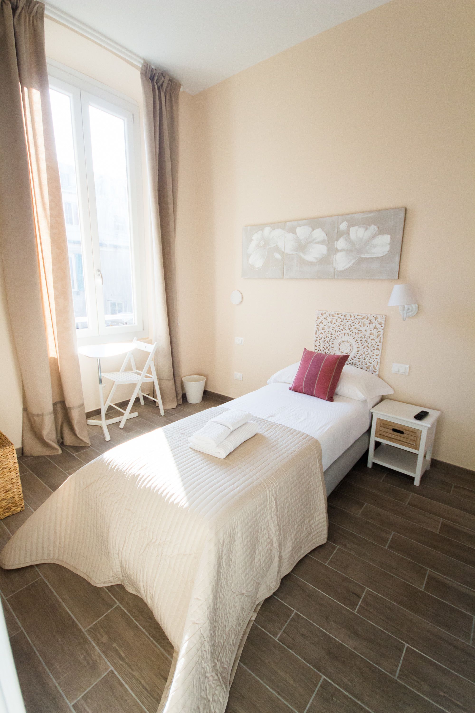 Un dormitorio con cama, mesita de noche, una silla, ventana con cortinas y un cuadro floral en la pared.