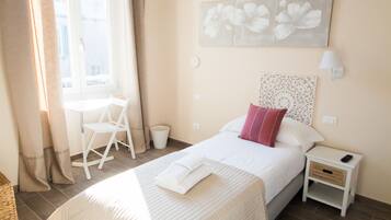 A bedroom with a bed, a nightstand, a chair, a window with curtains, and a wall with a floral painting.