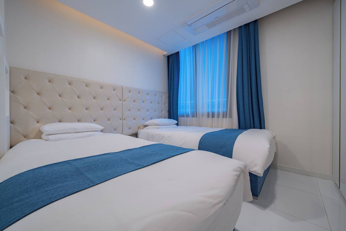 Quarto de hotel com duas camas, cabeceira estofada e uma janela grande com cortinas azuis.