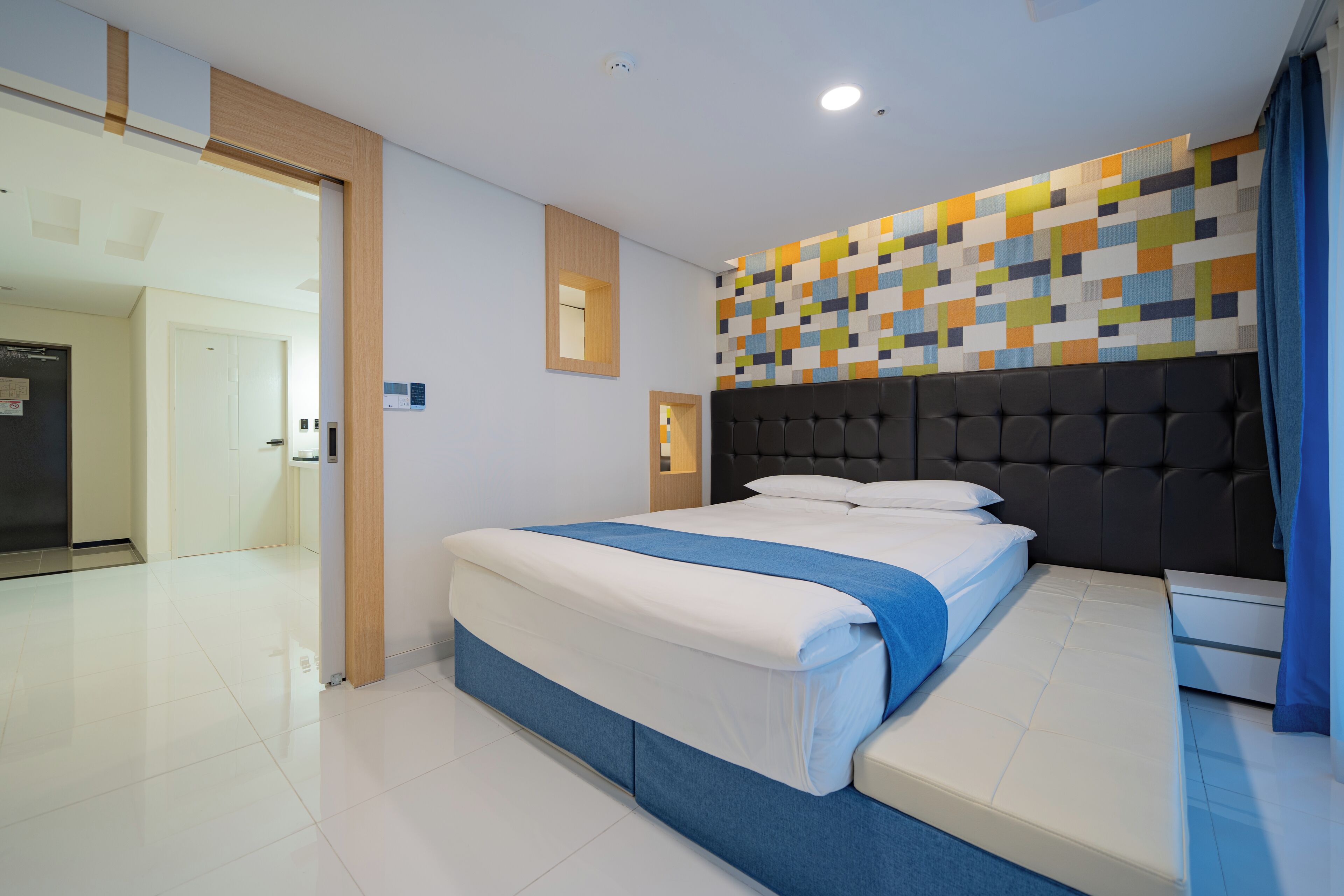 Ein modernes Hotelzimmer mit einem großen Bett, einem geometrisch gemusterten Kopfteil und einer hellen Deckenleuchte.