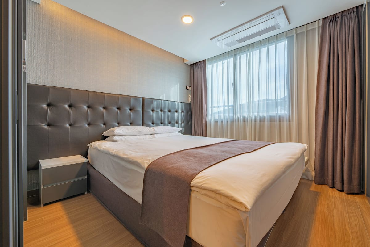 Quarto de hotel moderno com cabeceira estofada, uma cama de solteiro com roupa de cama branca, manta marrom, mesa de cabeceira com gavetas e uma janela grande com cortinas translúcidas.