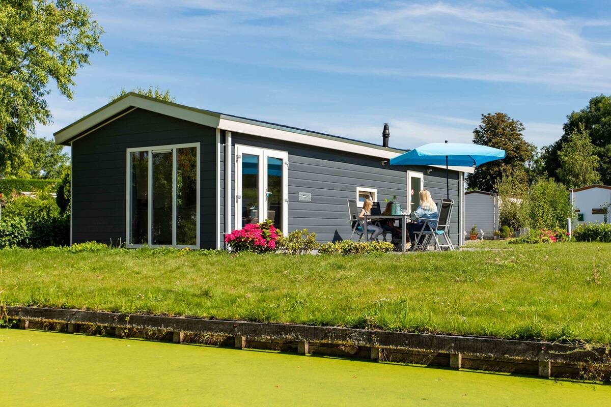 Ein Bungalow mit einer überdachten Terrasse, einem Eingang durch eine Glastür und einem kleinen Garten mit rosa Blumen.