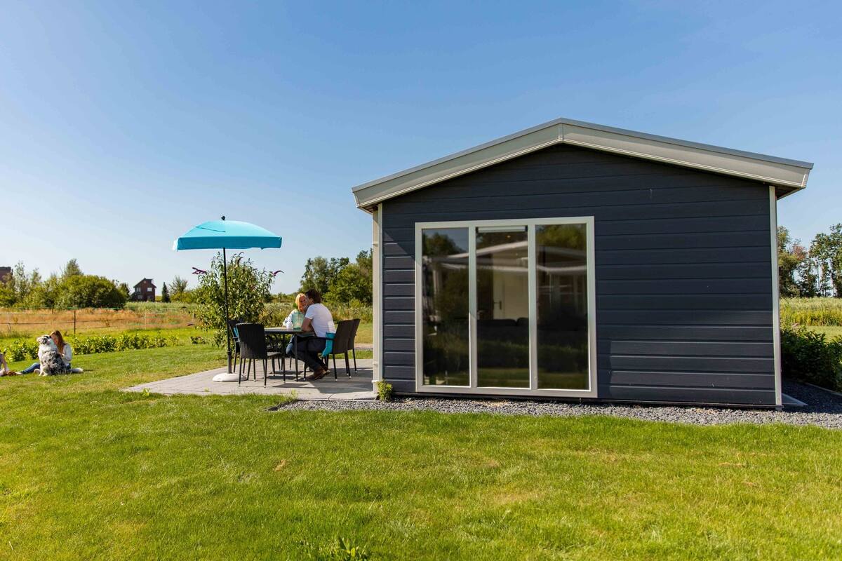 Ein kleines, dunkel gefärbtes Gebäude mit einer großen Schiebeglast ür, umgeben von einer gepflegten Rasenfläche und einer Terrasse mit Sitzgelegenheiten im Freien.