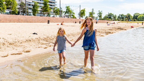 Two people walking hand in hand on a sandy beach with clear water, palm trees, and buildings in the background. - TopParken Parc de IJsselhoeve (Nieuwerkerk aan den IJssel)