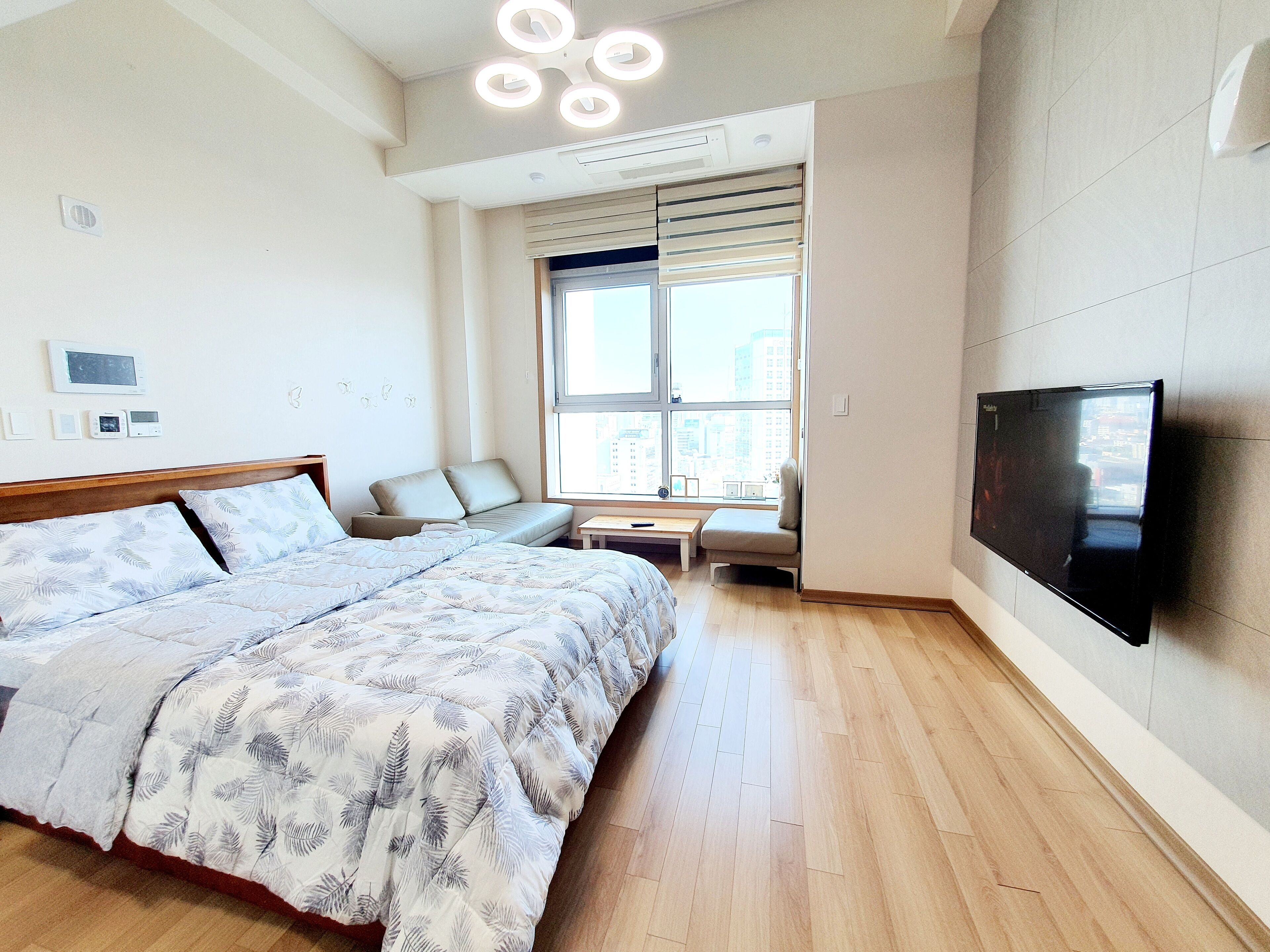 A modern bedroom with a large bed, a seating area, a TV, and a window with a city view.