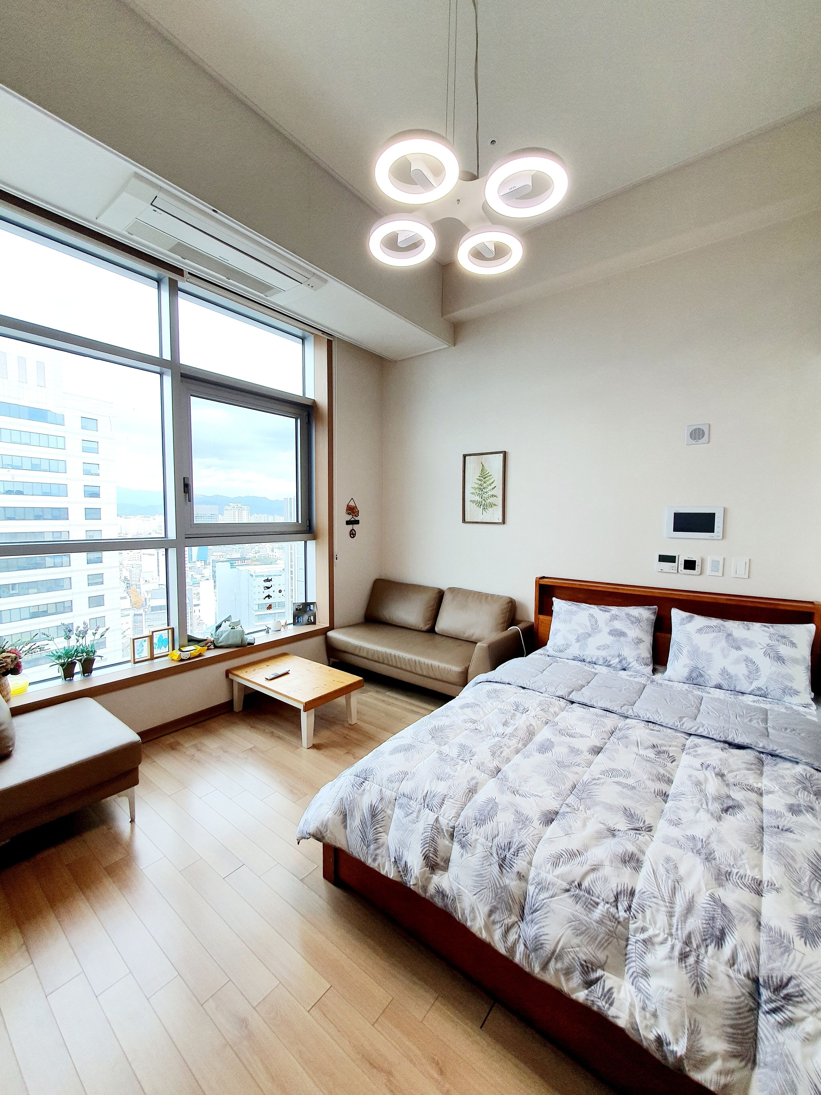 A modern bedroom with a large bed, a sofa, a small table, and a window with a city view.
