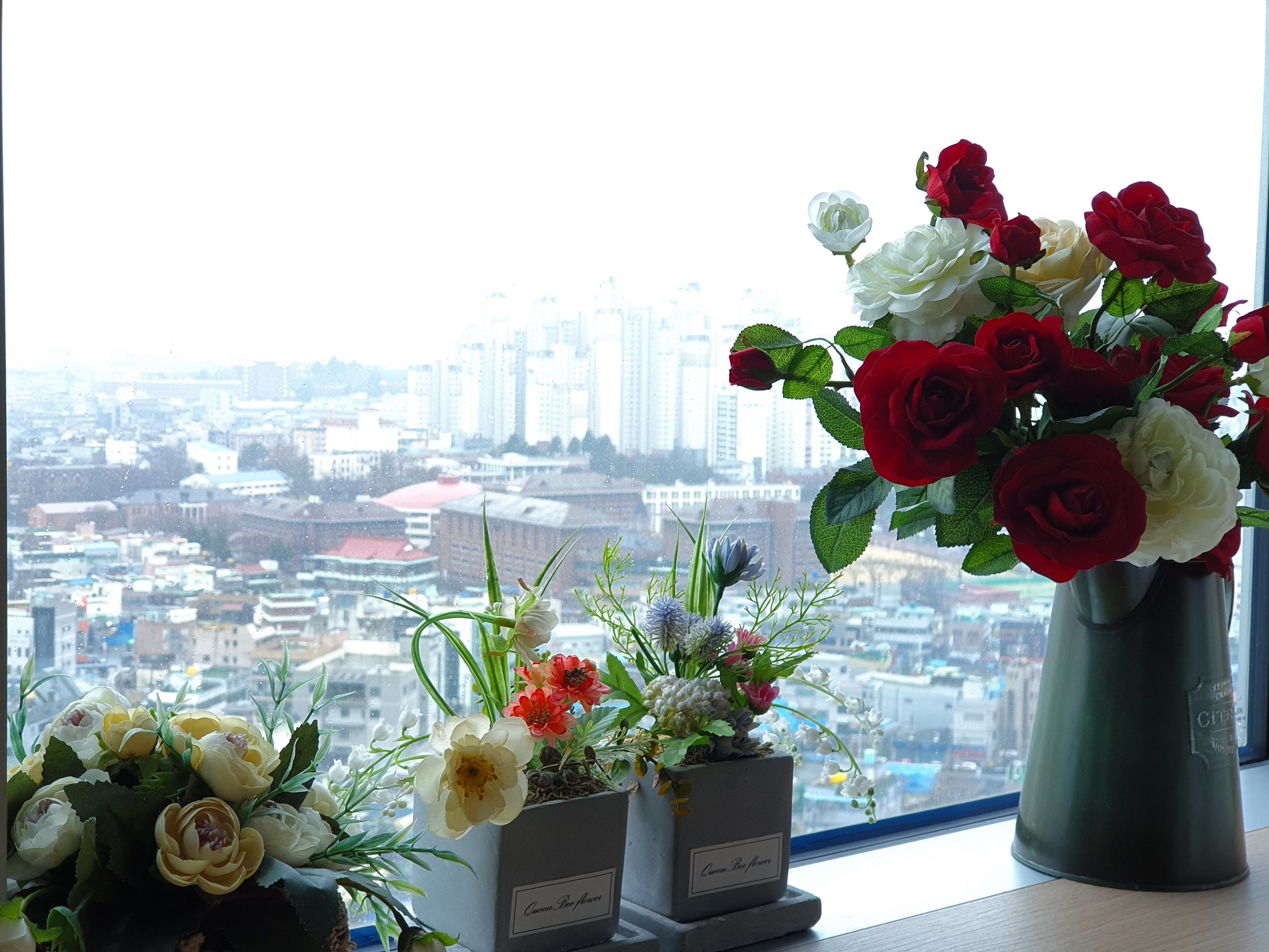 Una ventana con vistas a un paisaje urbano, un jarrón con flores rojas y blancas, y una maceta más pequeña con flores amarillas.