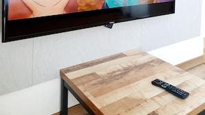 A flat-screen TV mounted on a wall with a remote control on a wooden table in front. - DG HOUSE (Daegu)