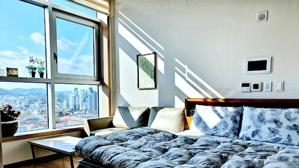 A bedroom with a large window, a bed with a leaf-patterned duvet, a bedside table, and a chair. - DG HOUSE (Daegu)