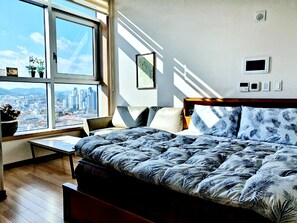 A bedroom with a large window, a bed with a leaf-patterned duvet, a bedside table, and a chair.