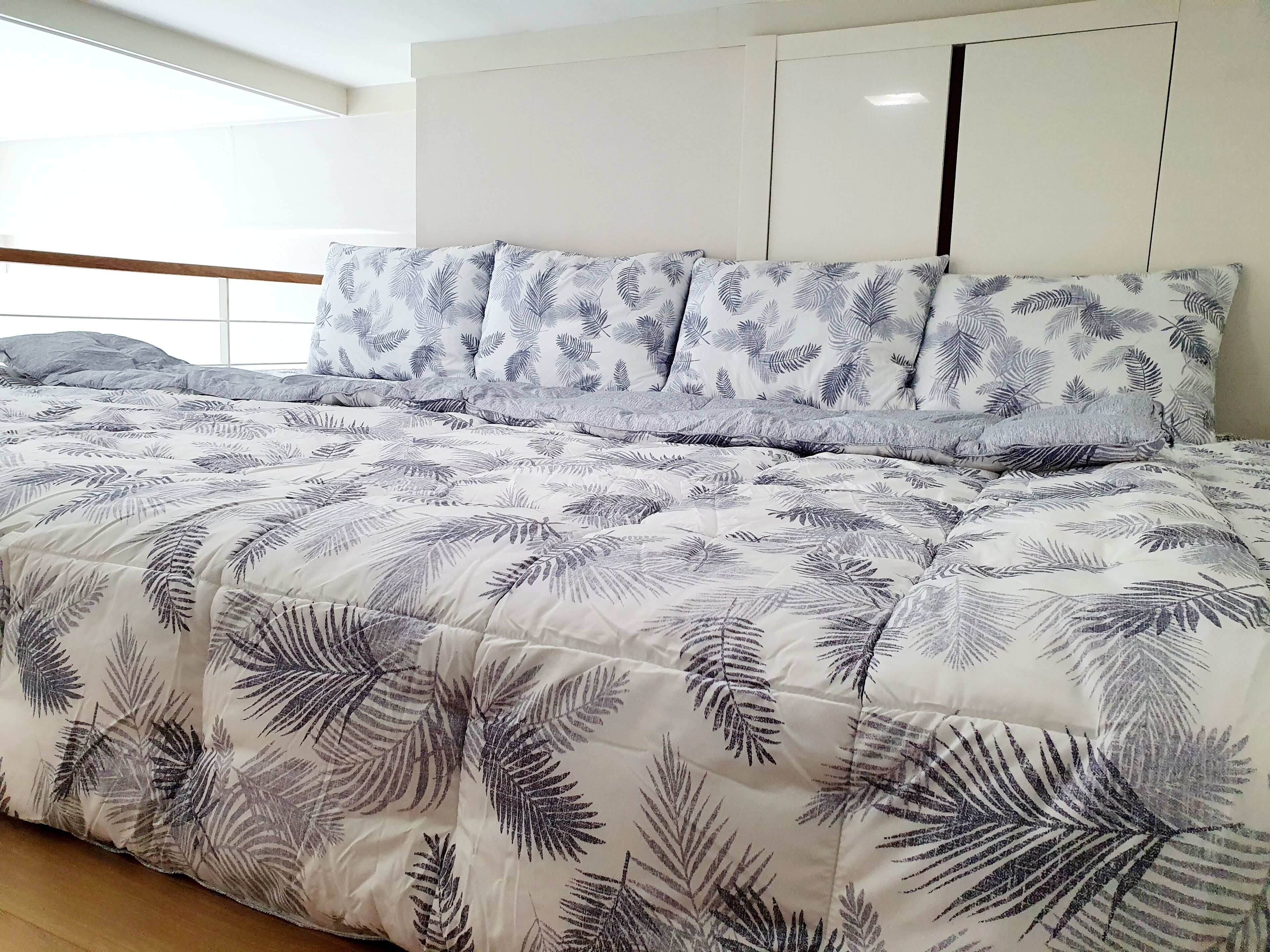 A bed with a leaf-patterned duvet and pillows, a wooden headboard, and a window with a view of the outdoors.