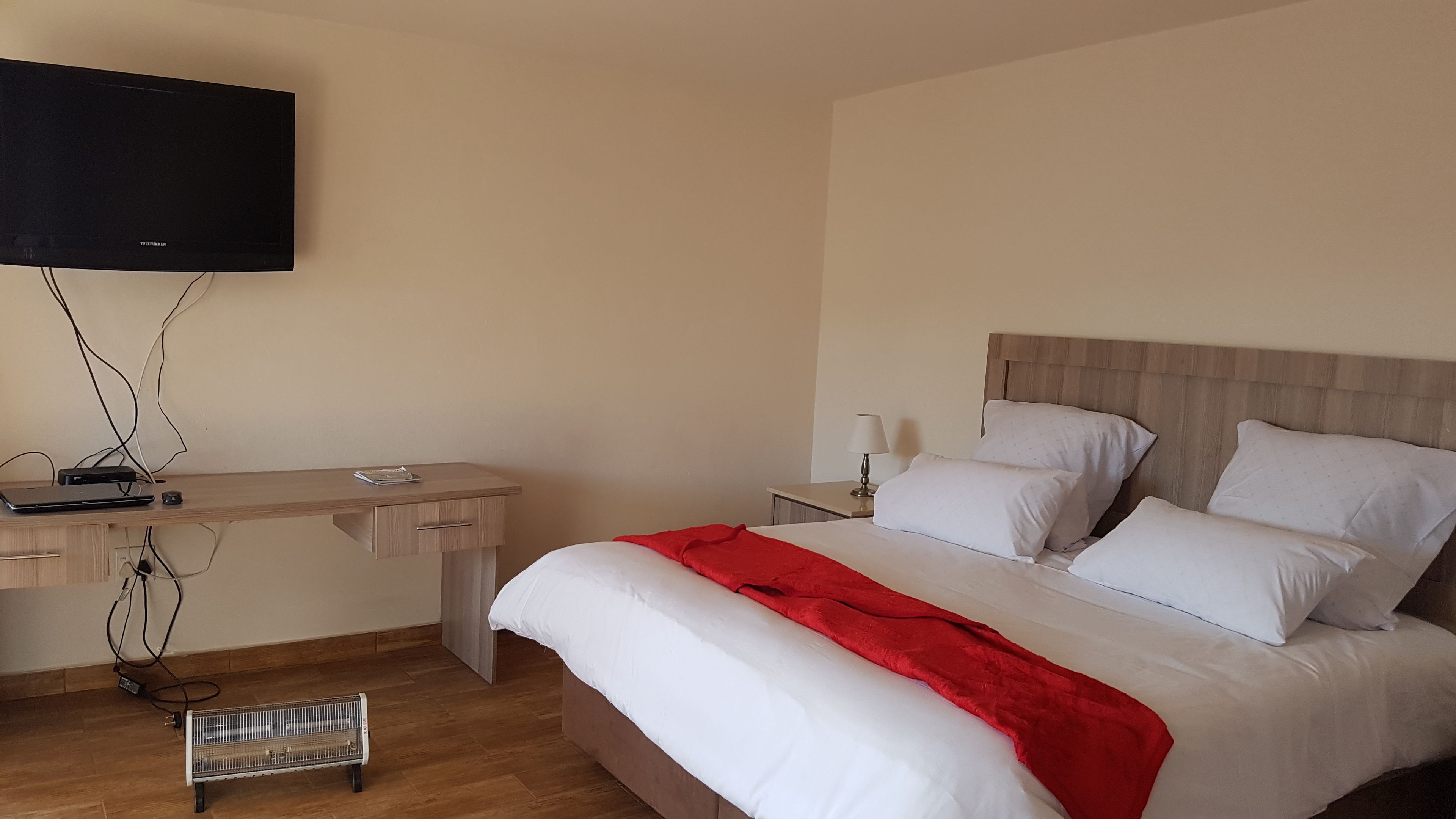 A hotel room with a bed, a desk, a television mounted on the wall, and a heater on the floor.