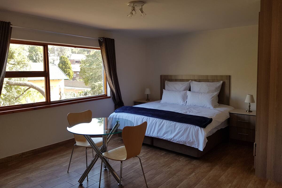 A bedroom with a bed, bedside tables, a window with curtains, a table with chairs, and a ceiling light.
