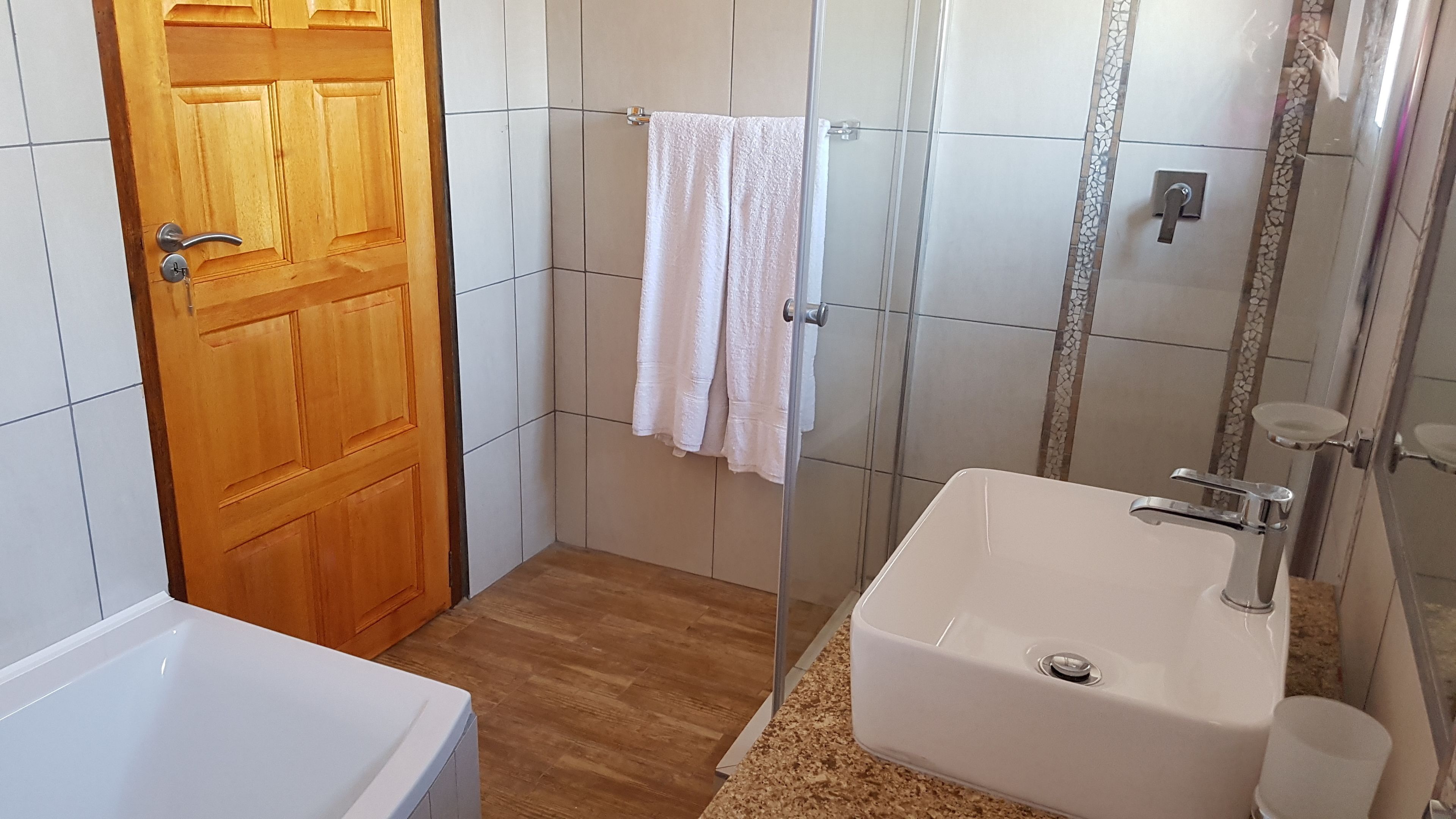 A bathroom with a wooden door, white tiles, a white sink, and a shower area.