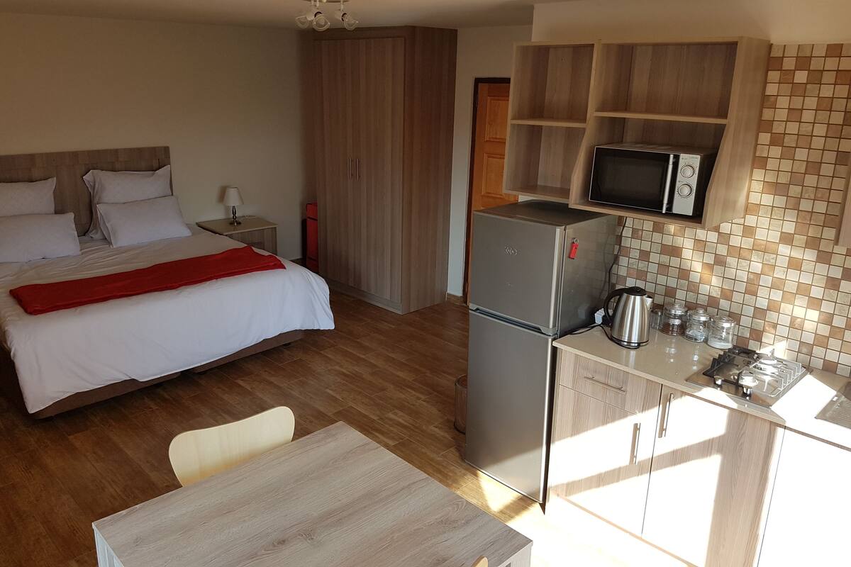 A compact hotel room with a bed, a small table, a kitchenette with a refrigerator and microwave, and a wooden wardrobe.