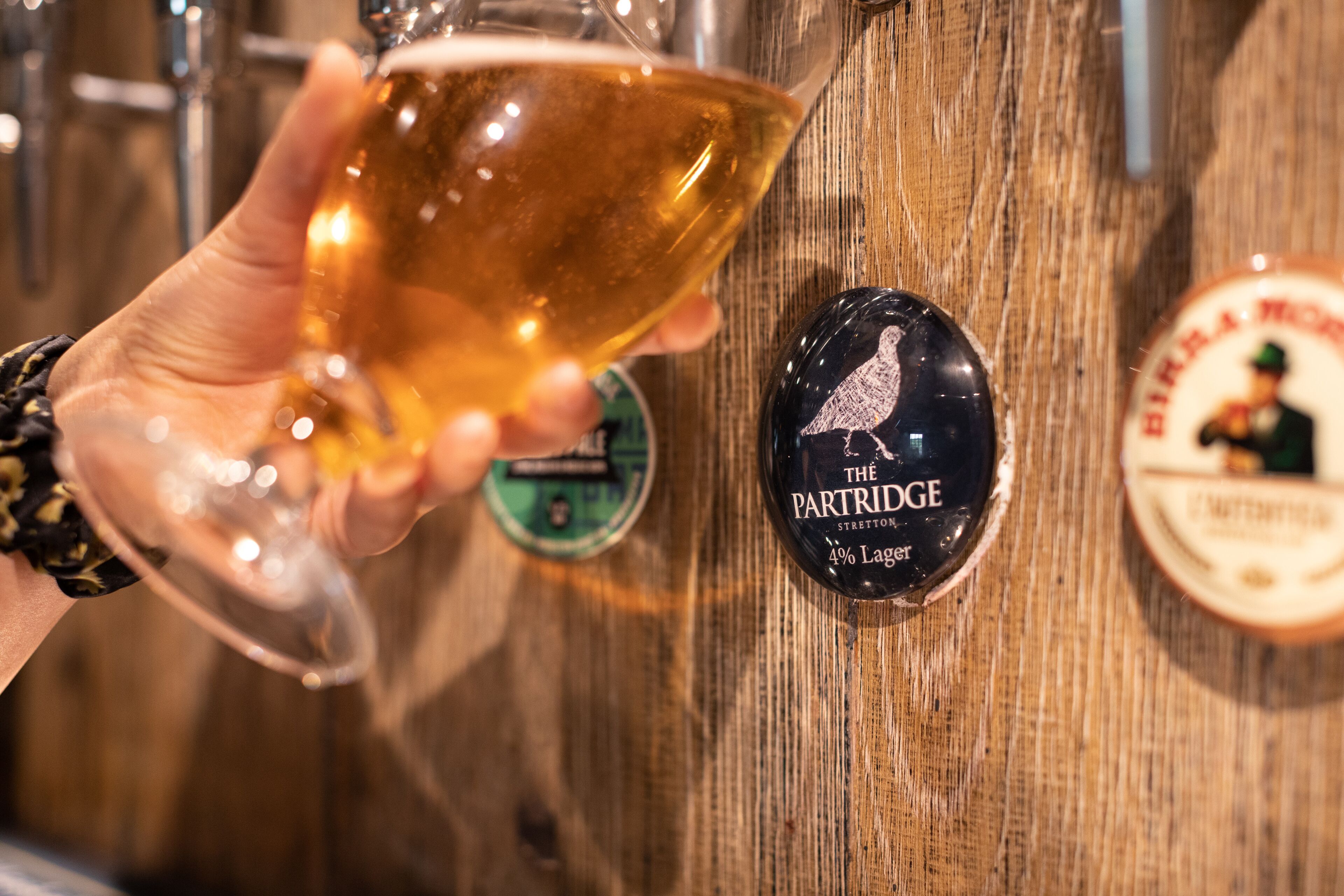 A hand holding a glass of beer with a wooden background and a label reading "The Partridge 4% Lager".