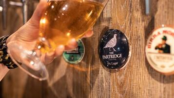 A hand holding a glass of beer with a wooden background and a label reading "The Partridge 4% Lager".