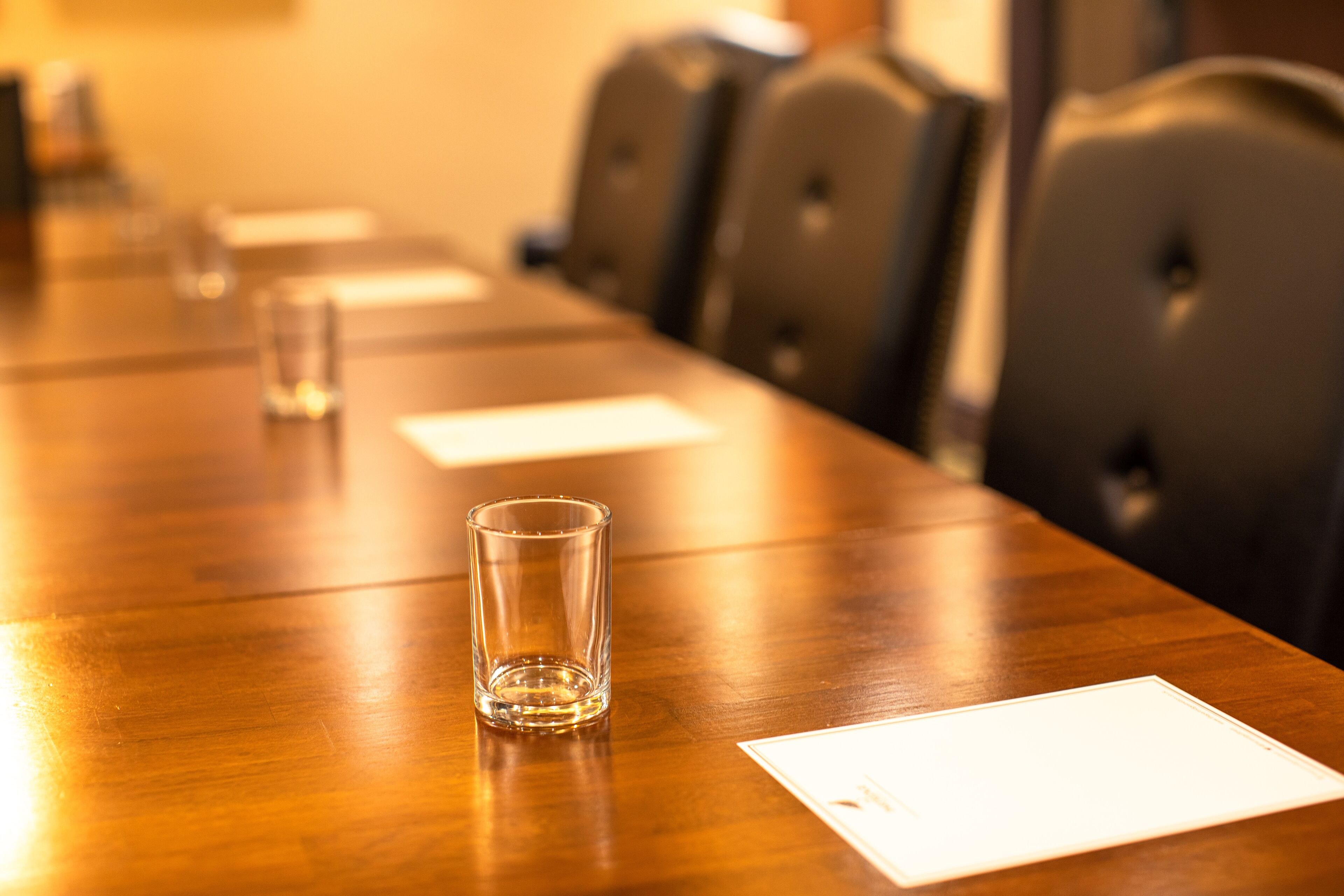 A conference table with chairs, a glass, and a white sheet of paper.