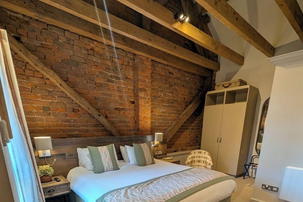 A bedroom with a large bed, a wooden ceiling, a brick wall, a wardrobe, and a nightstand with a lamp.