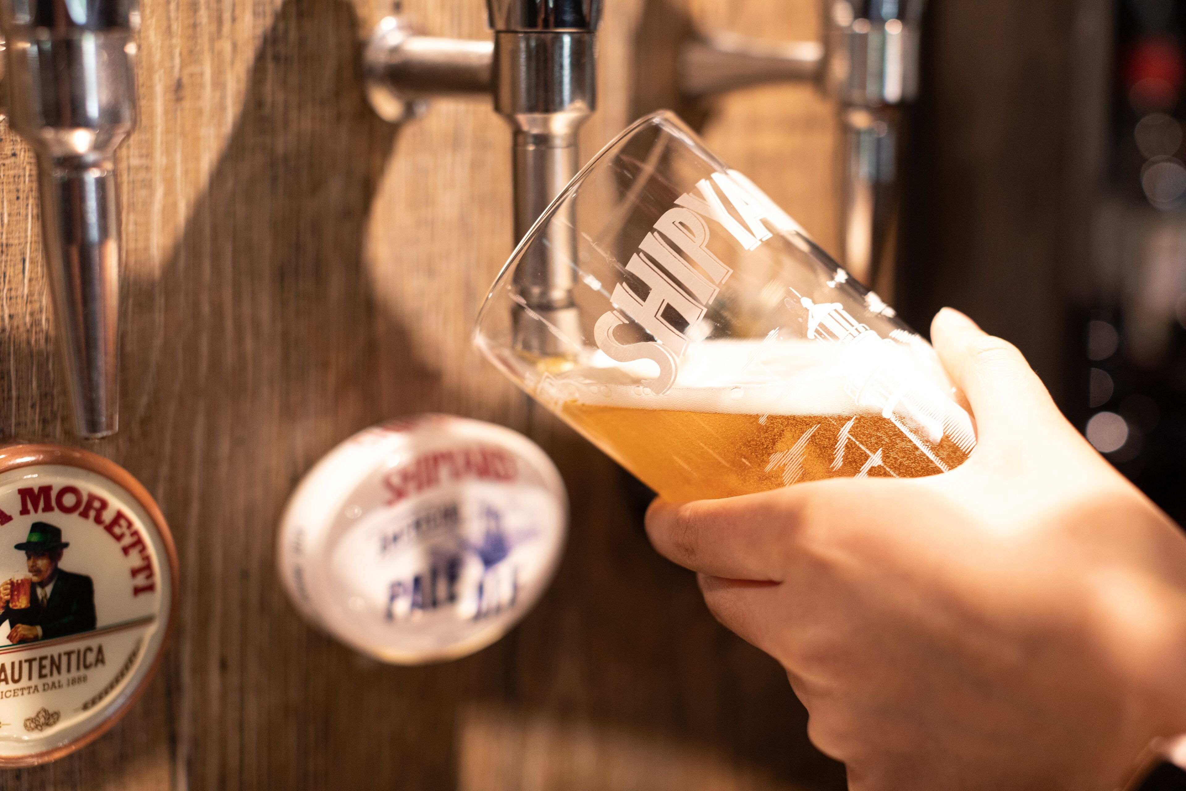 A hand holding a glass with "SEMPRE" embossed on it, positioned in front of a beer tap.