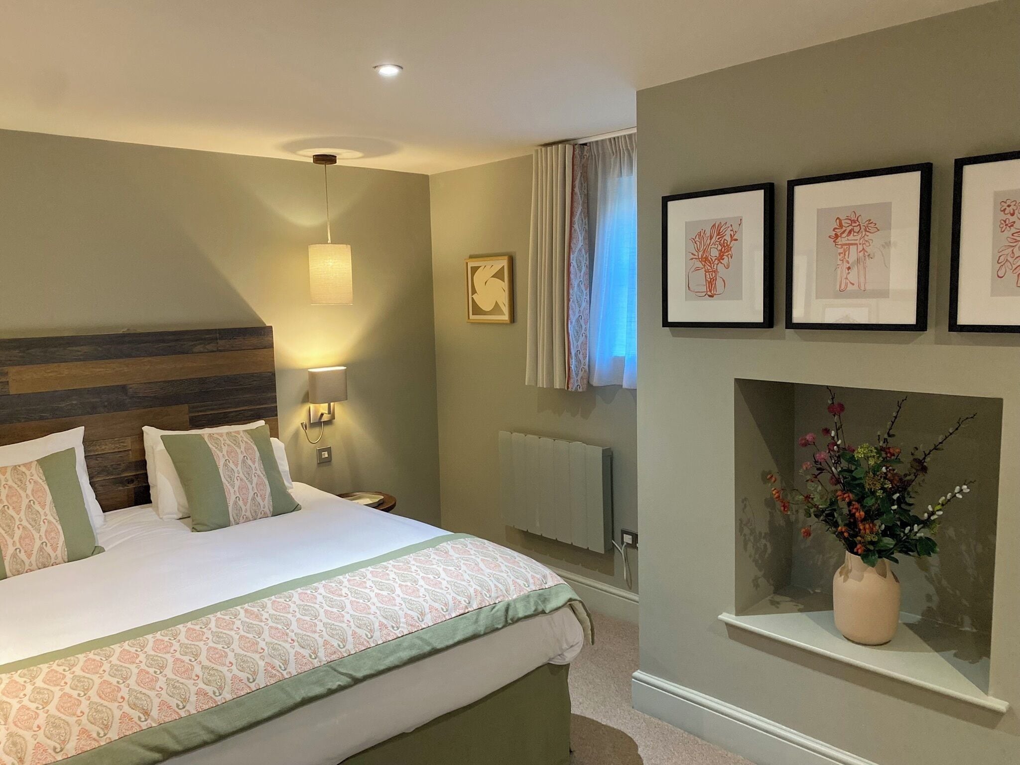 A hotel room with a bed, bedside lamp, framed artwork, and a vase with flowers.