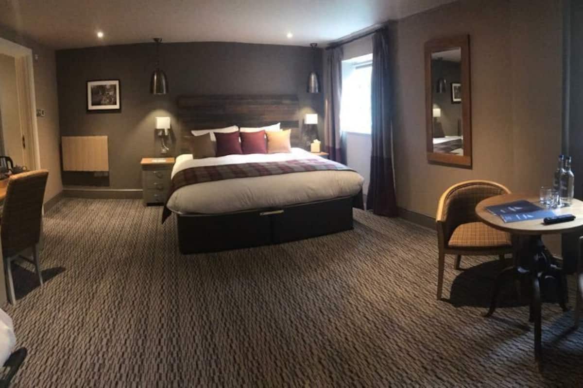 A hotel room with a large bed, a desk with a chair, a small table with a chair, and a window with curtains.