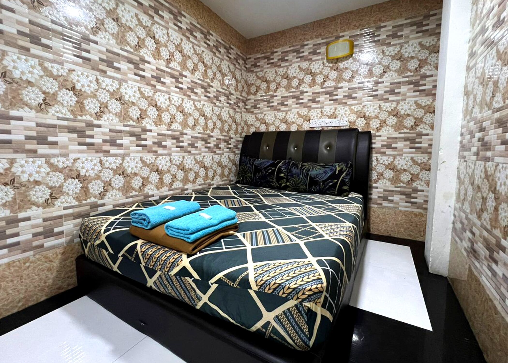 A small, neatly arranged bedroom with a patterned wall, a bed with a geometric patterned bedspread, and two folded towels on the bed.
