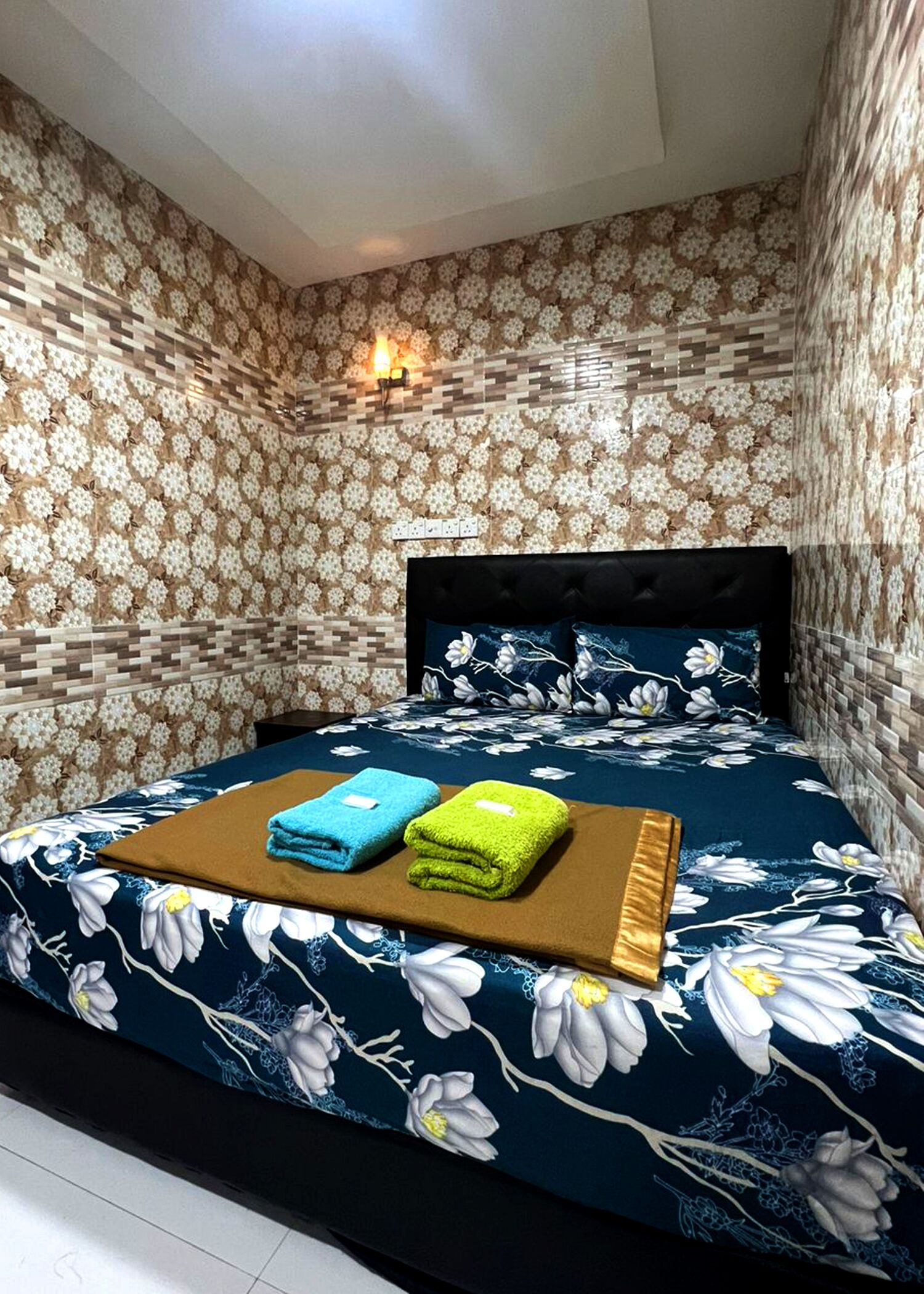 Un dormitorio con una colcha de estampado floral, un cabecero oscuro y una lámpara fijada a la pared.