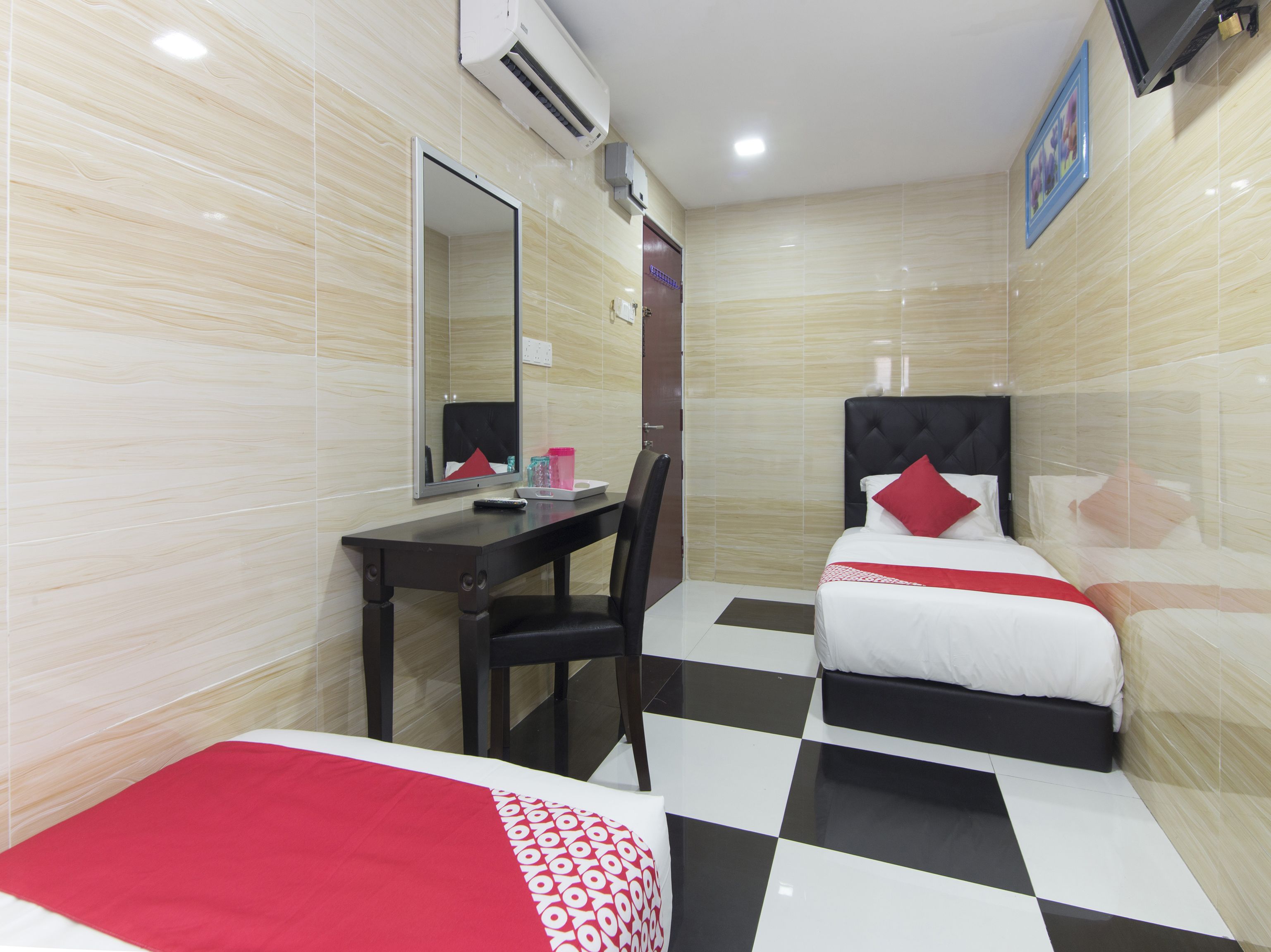 Una habitación de hotel con piso a cuadros, un escritorio con una silla, una cama con un almohón rojo y un espejo grande en la pared.