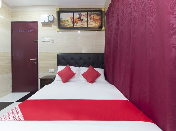 Een hotelkamer met een bed, een rood-witte spreideken, twee rode kussens, een zwart hoofdeinde, een houten nachtkastje, een rode deur, een schilderij aan de muur en een gordijn met een patroon.
