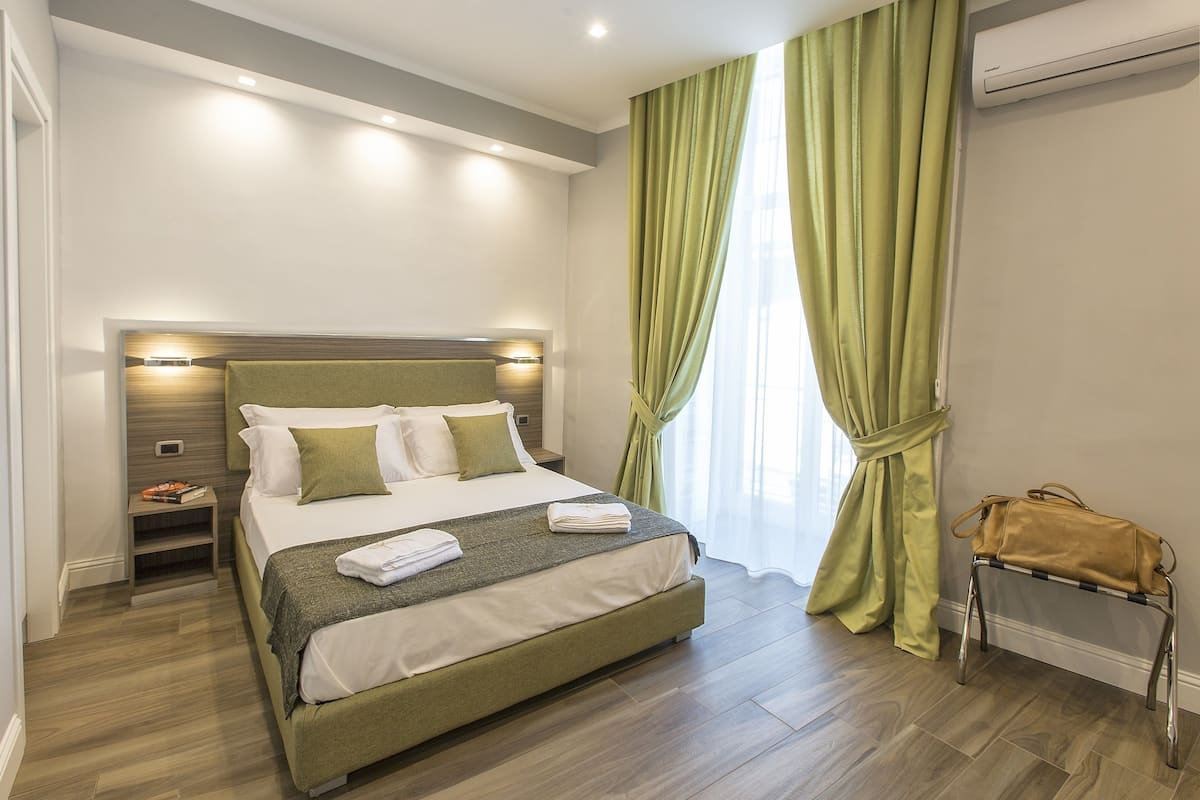 A neatly made bed with a green headboard, a bedside table with a lamp, and a window with green curtains.